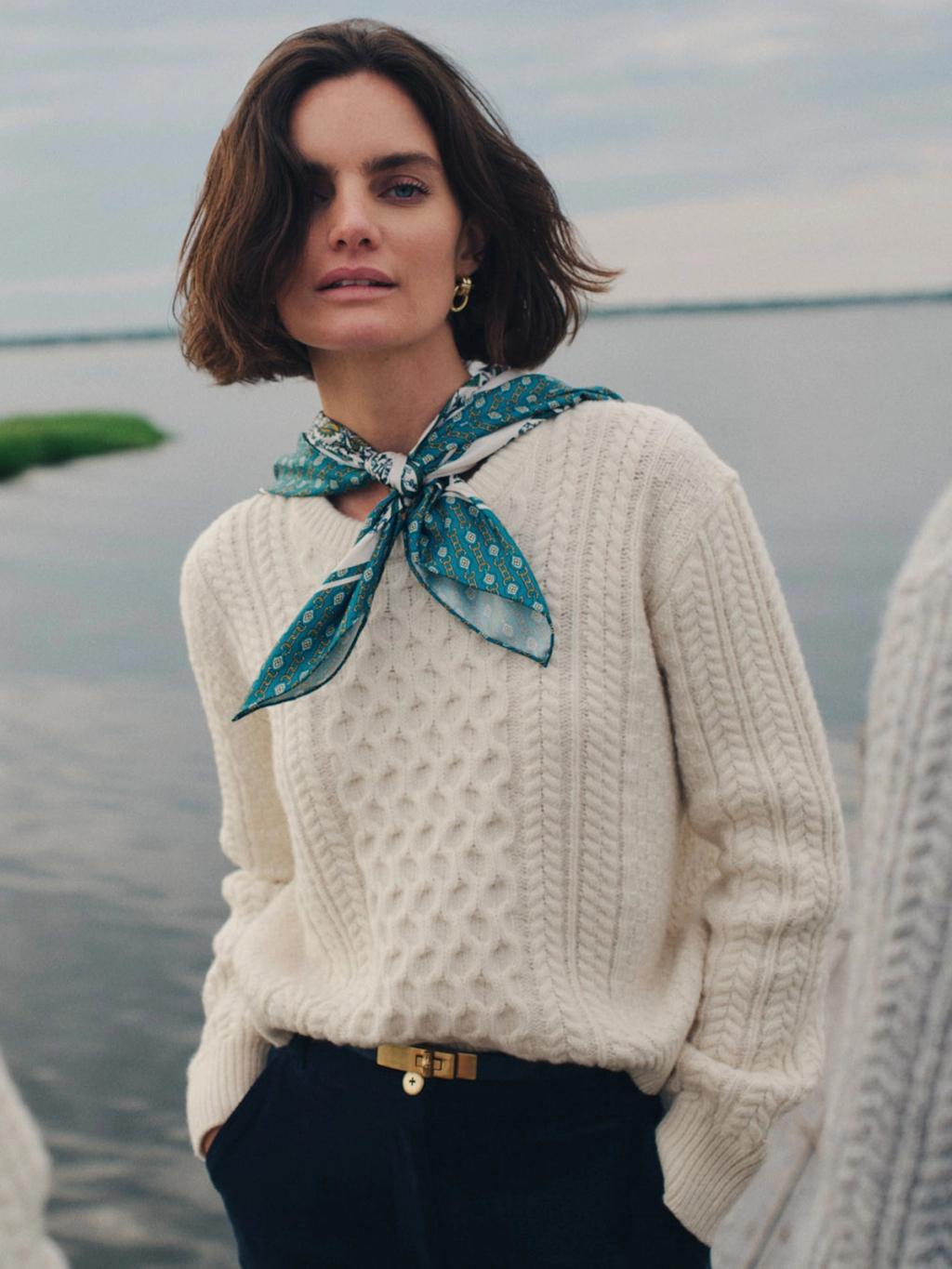 A woman in a cream cable-knit sweater and blue patterned scarf looks at the camera by a body of water.