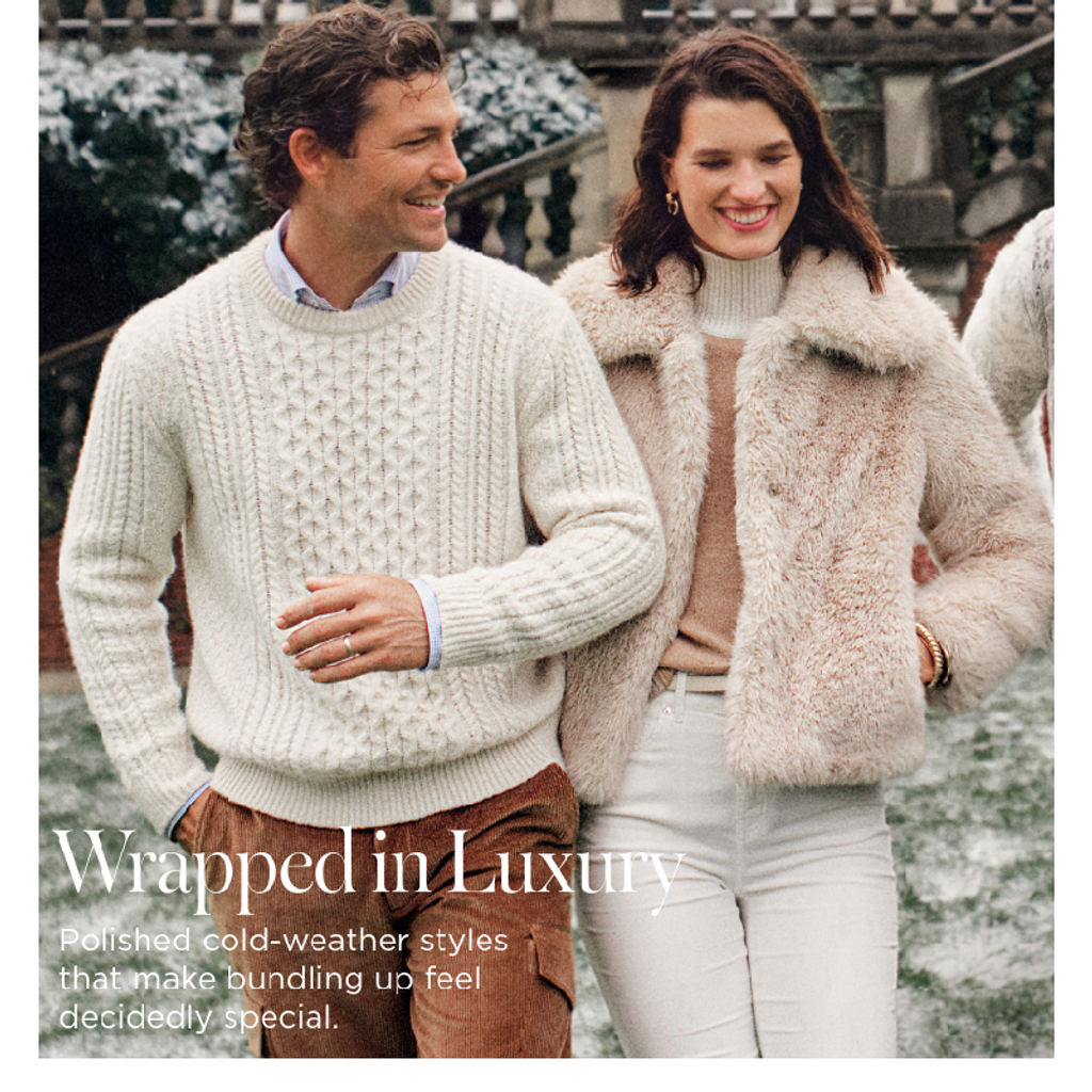 A smiling man in a cream cable-knit sweater and a woman in a light beige faux fur coat walk in a winter setting.