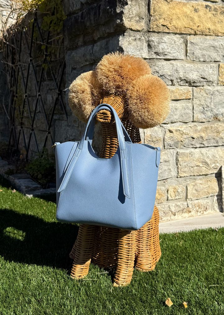 A light blue handbag rests on a wicker dog-shaped display stand in a grassy outdoor setting with a stone wall in the background.