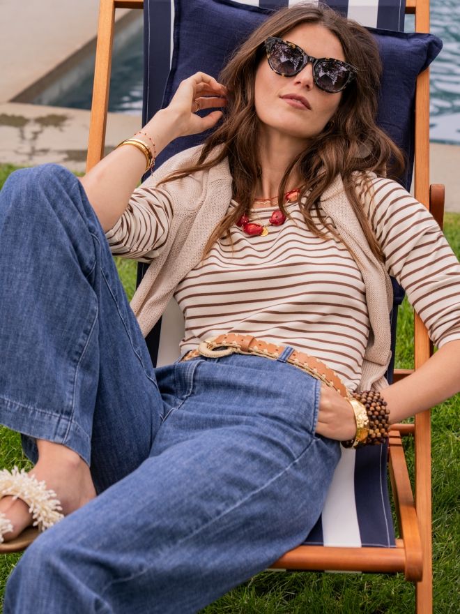 woman outside leaning back while seated in a blue and white striped lounge beach chair.  She is wearing brown and cream striped cotton tee and medium indigo jeans
