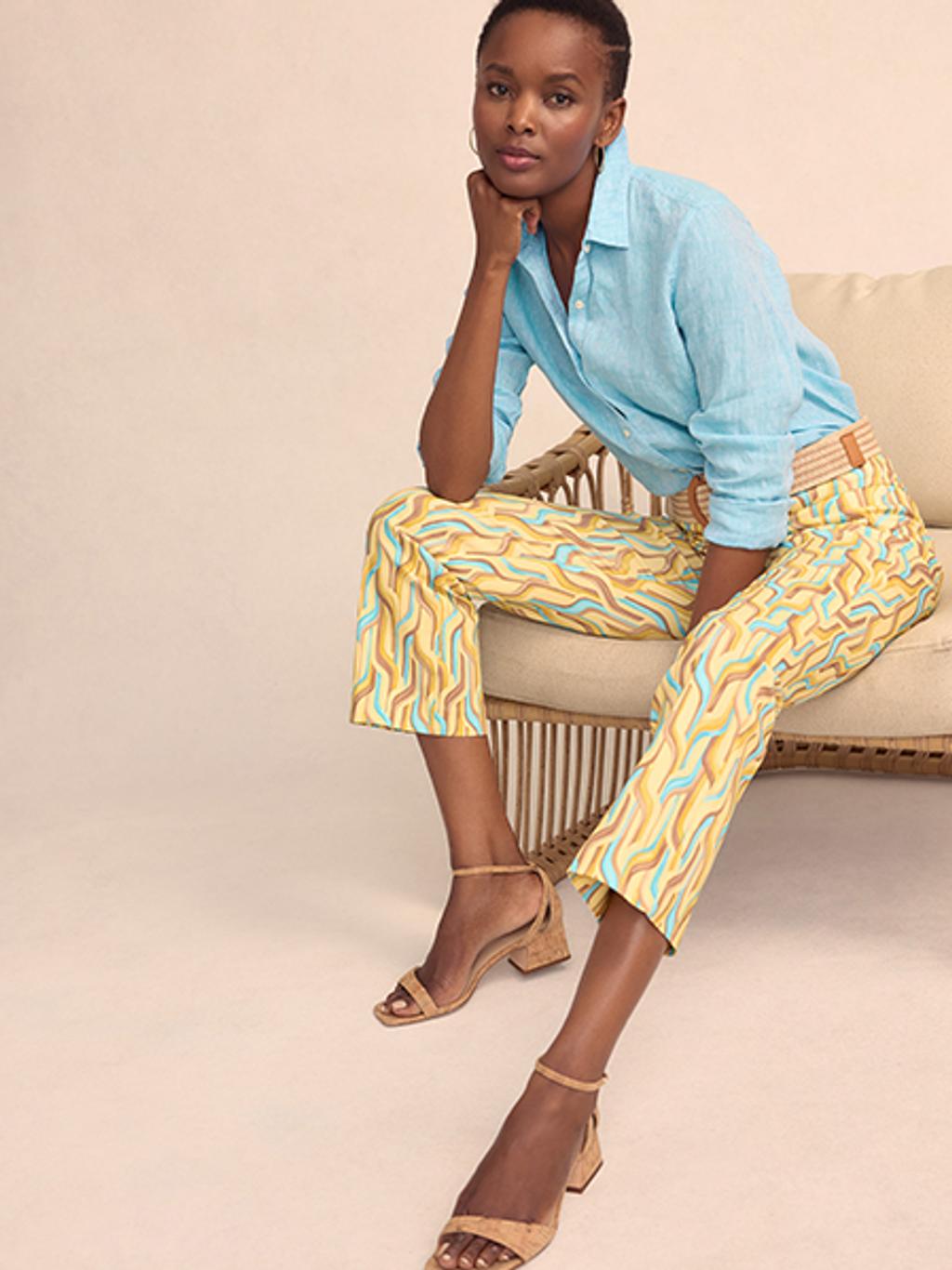 Woman sitting while wearing a light blue linen button up shit and patterned capri pants, paired with tan block heel sandals.