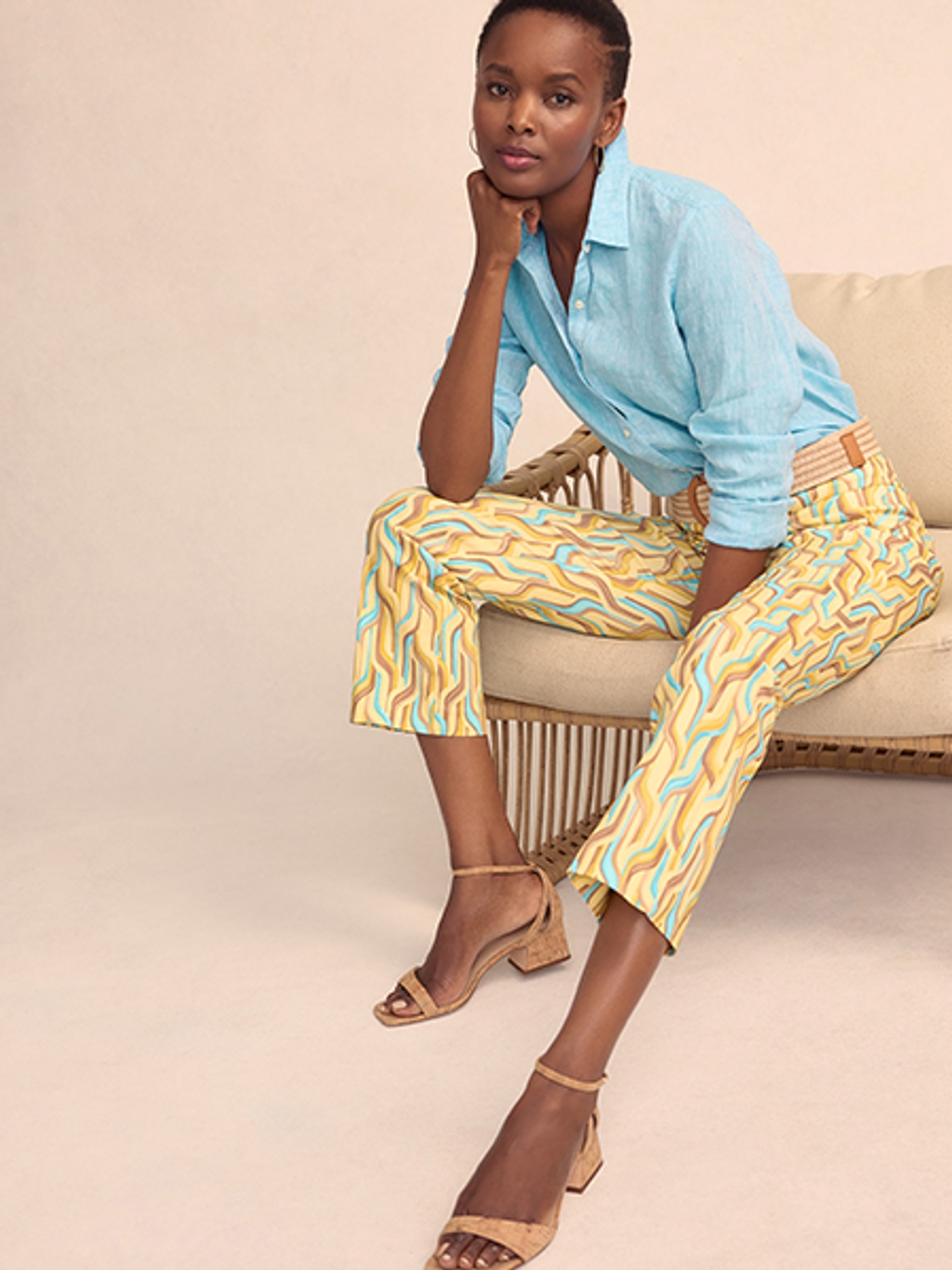 Woman sitting while wearing a light blue linen button up shit and patterned capri pants, paired with tan block heel sandals.
