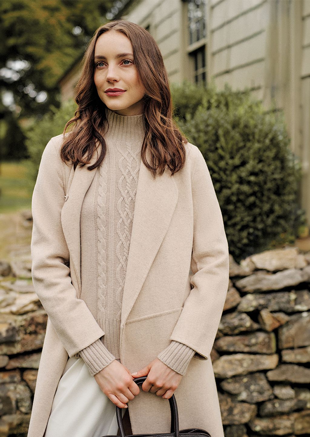 Woman standing outside in cream jacket, cream sweater, and white pants.