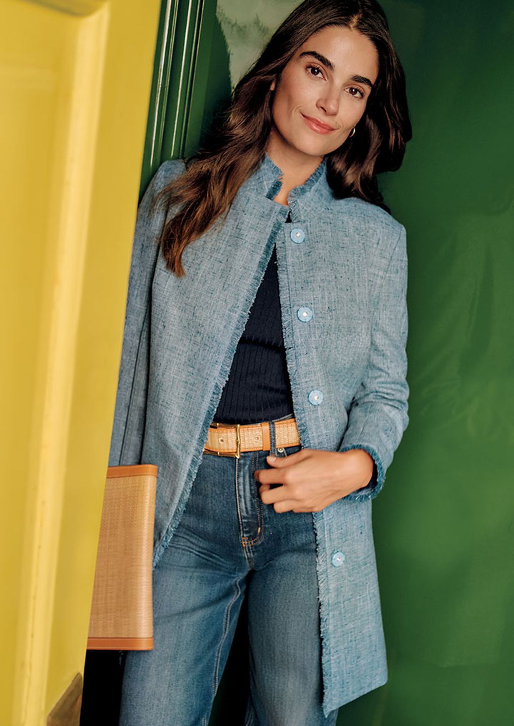 A woman in a blue coat, navy sweater, jeans and a grasscloth belt and clutch.