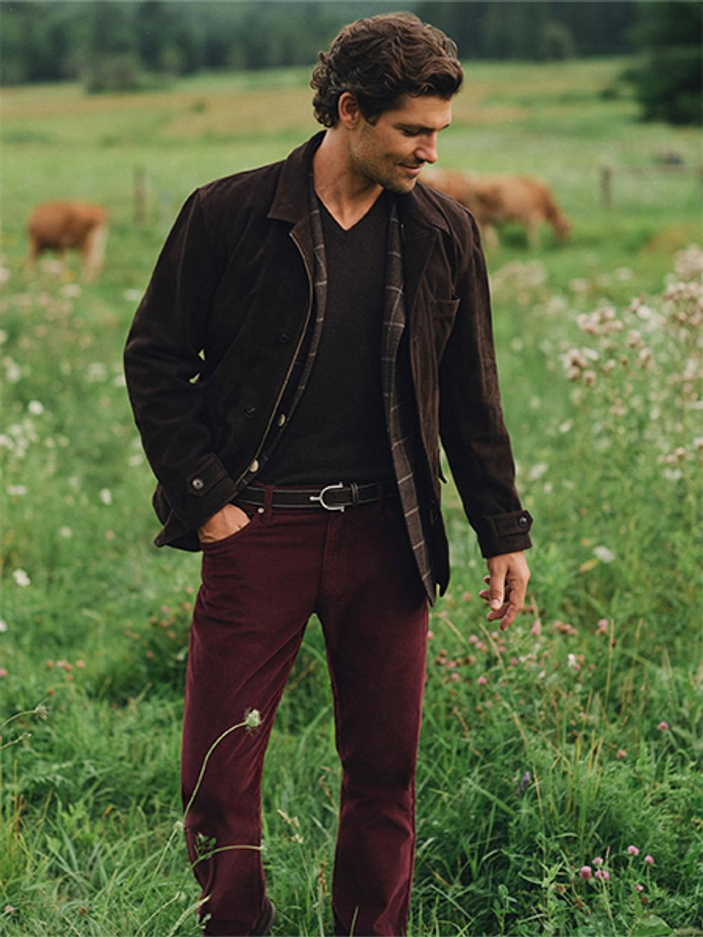 A man in a brown suede jacket and burgundy corduroy pants stands in a green field with cows.
