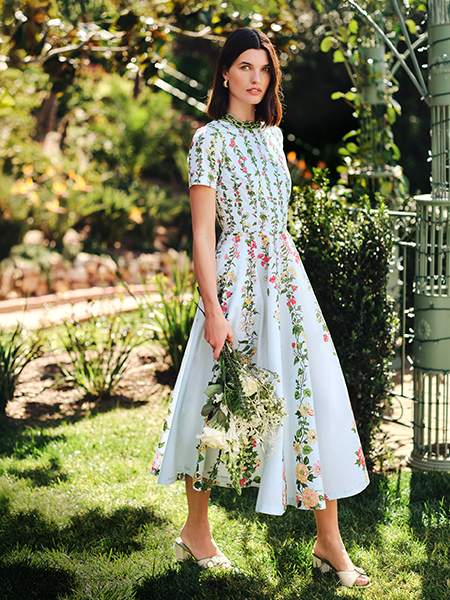 A woman in a light blue floral dress, holding white flowers, stands in a lush garden.