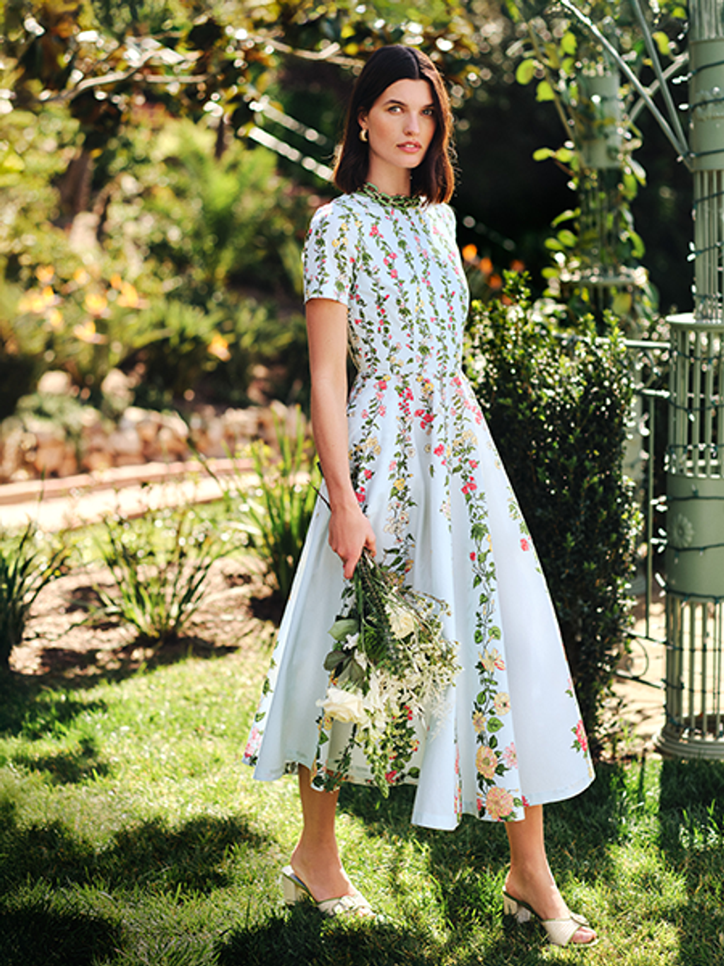 A woman in a light blue floral dress, holding white flowers, stands in a lush garden.