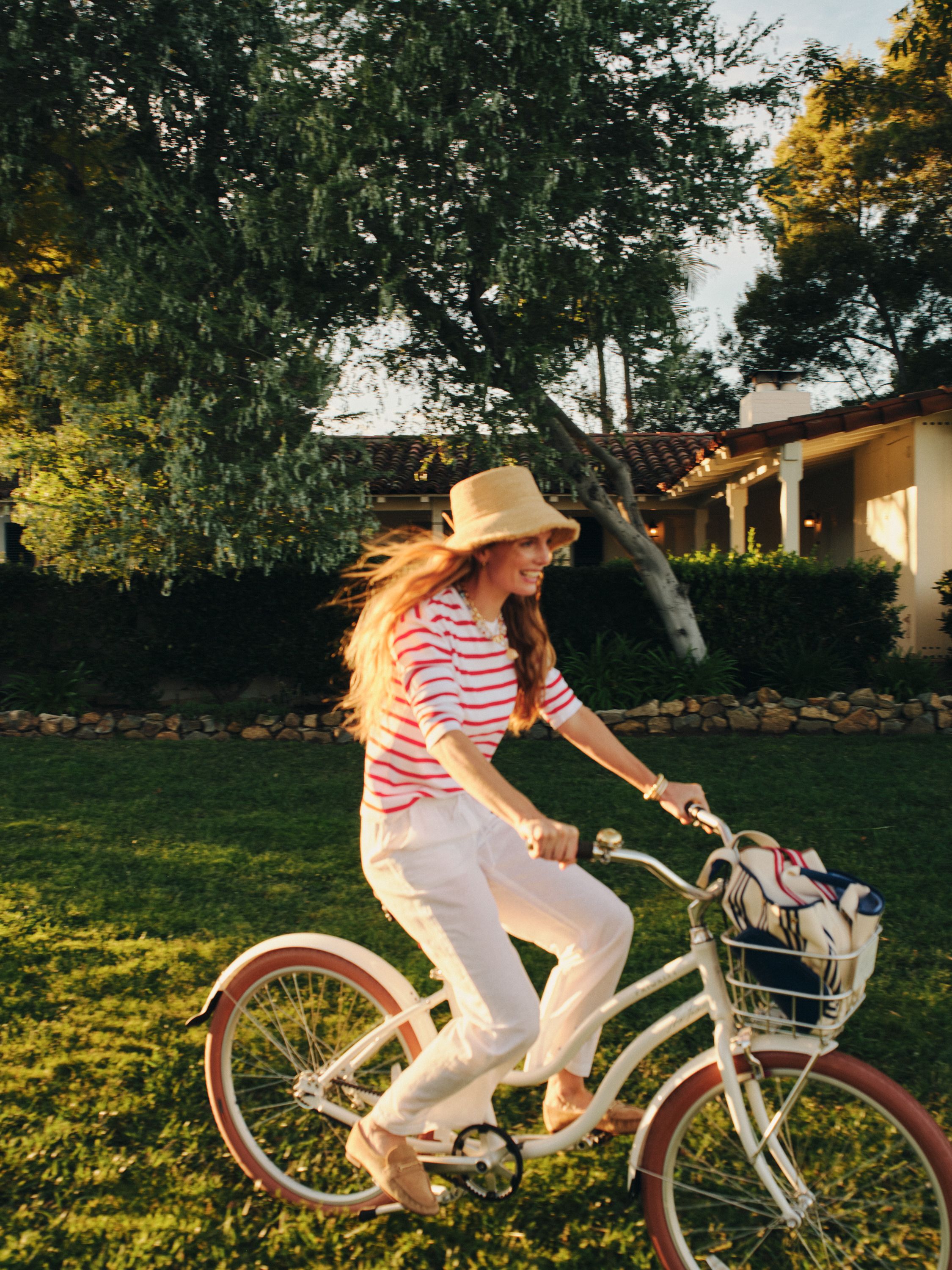 Woman in straw hat and striped shirt smiling while riding a white bicycle on a sunny lawn.