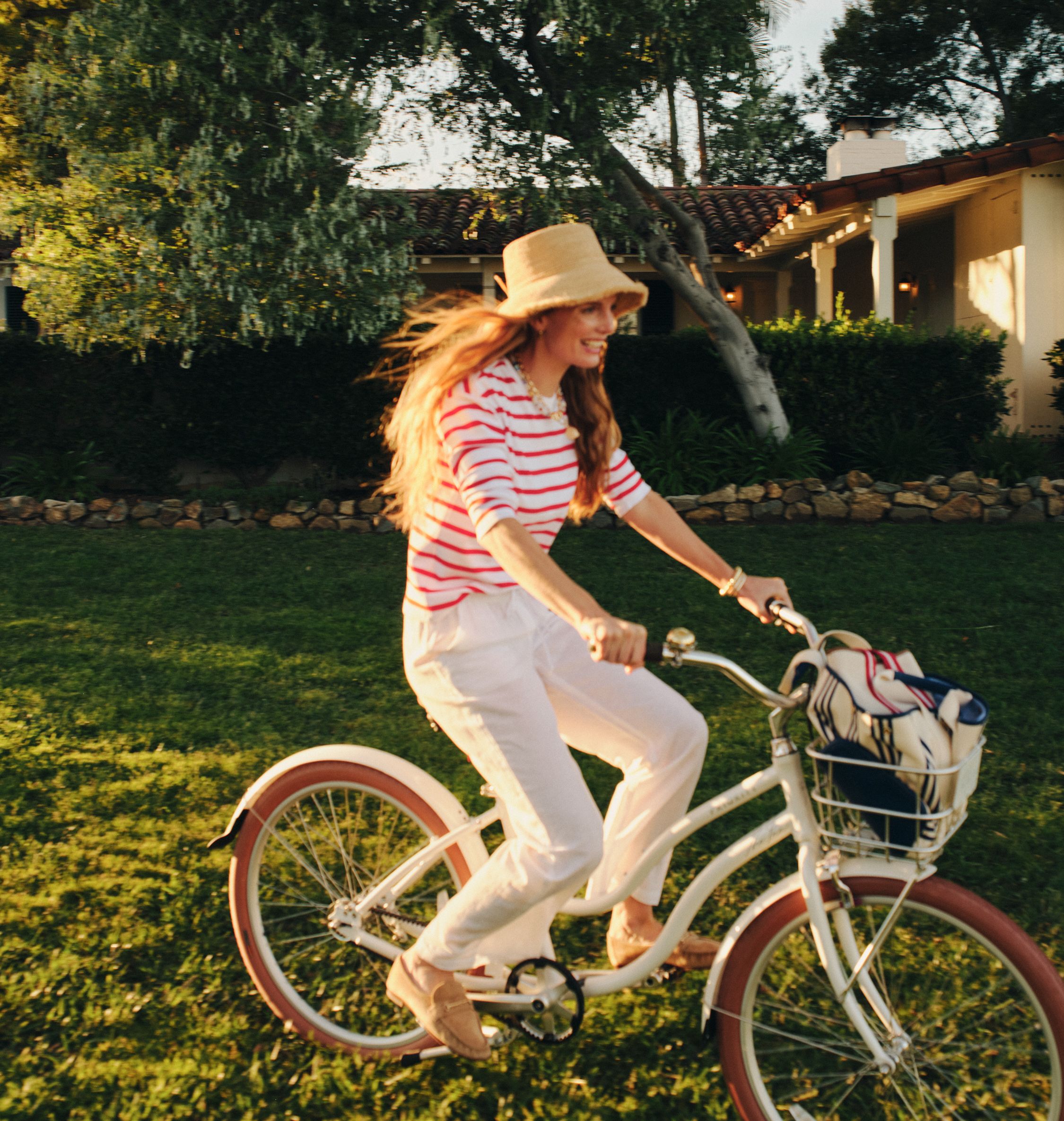 Woman in straw hat and striped shirt smiling while riding a white bicycle on a sunny lawn.