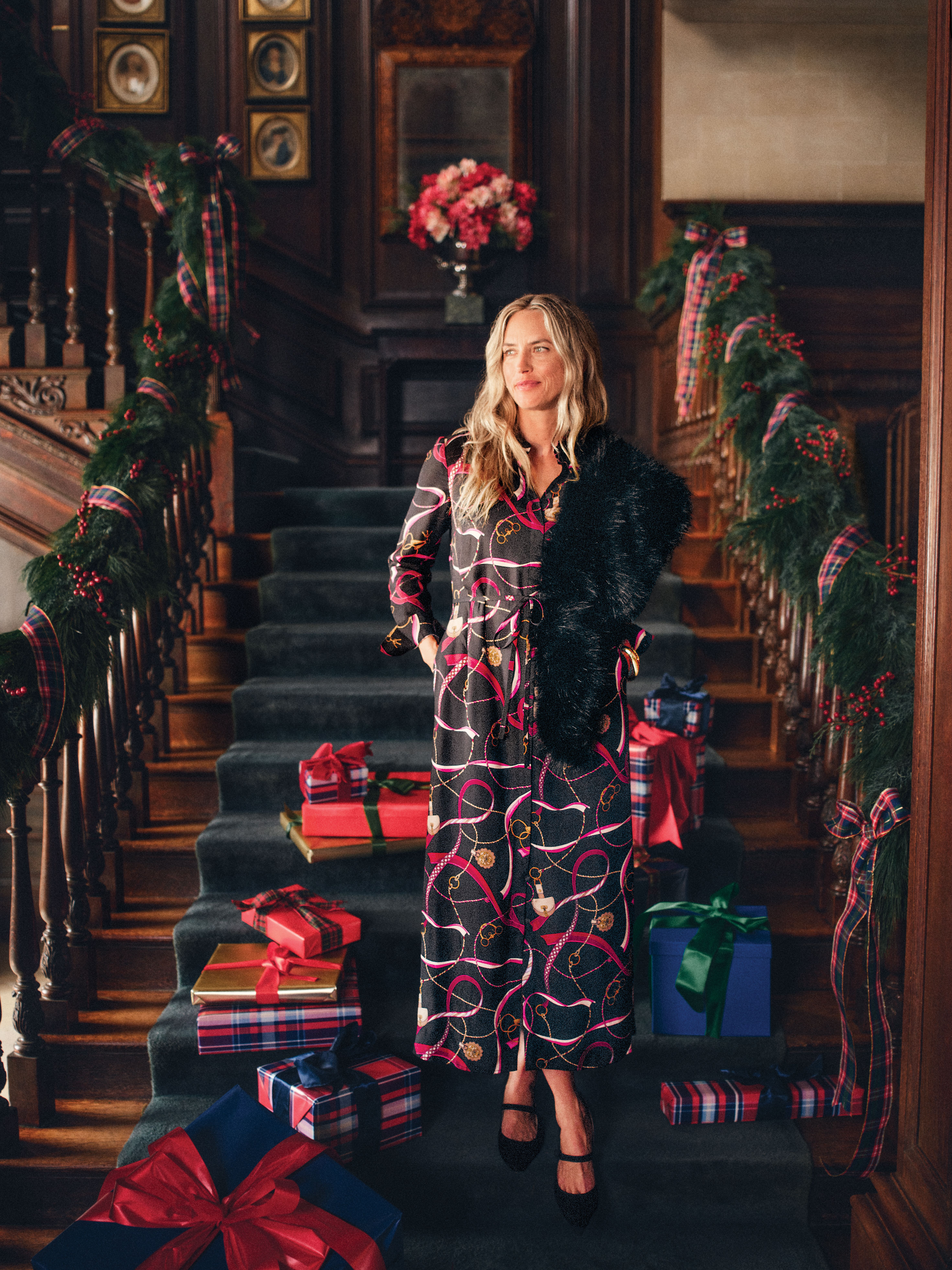 A woman in a patterned dress and black fur stole on a holiday-decorated grand staircase with wrapped gifts.