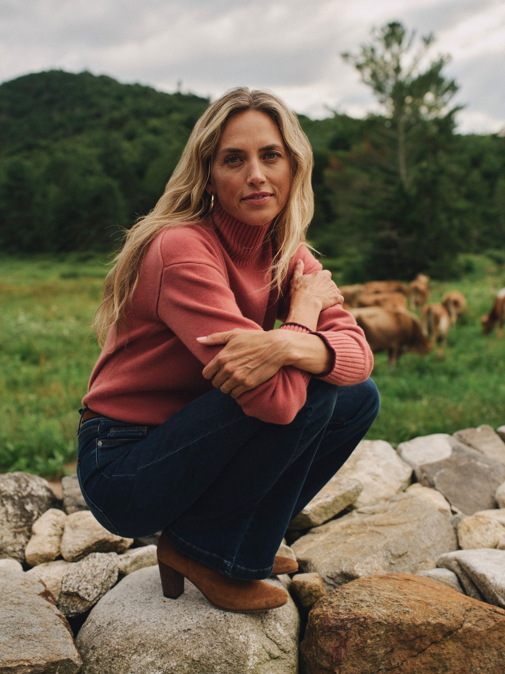 A woman in a pink turtleneck and dark jeans crouches on a stone wall, with a green hillside and cows in the background.