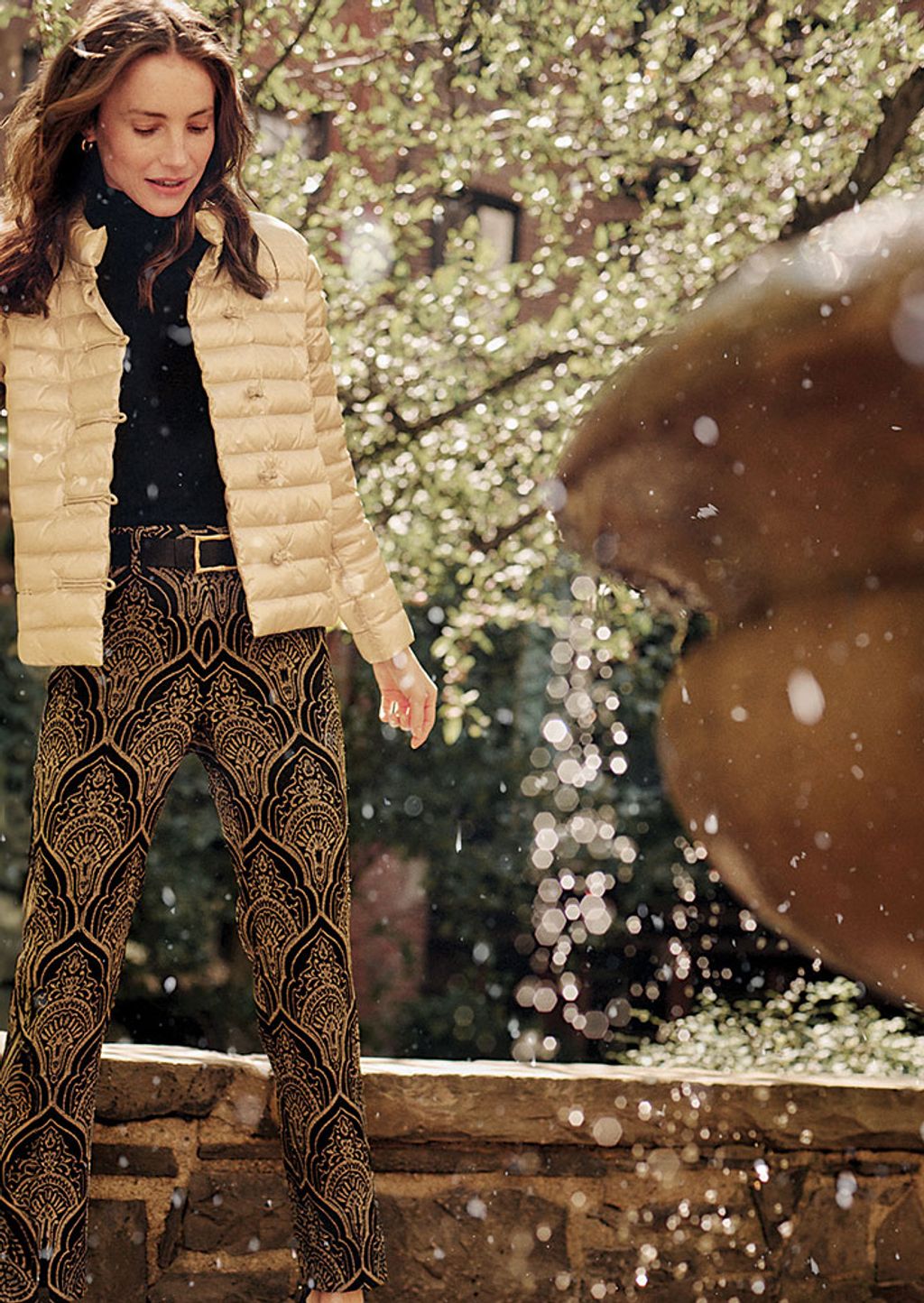 A woman wearing a gold puffer jacket, black turtleneck, and black and gold jacquard pants.