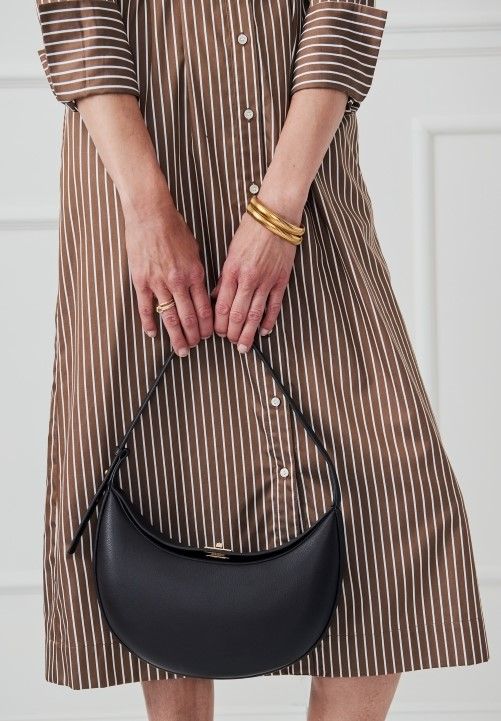 cropped image of woman holding brown leather half moon shoulder bag with turnlock closure, standing in front of white studio backdrop