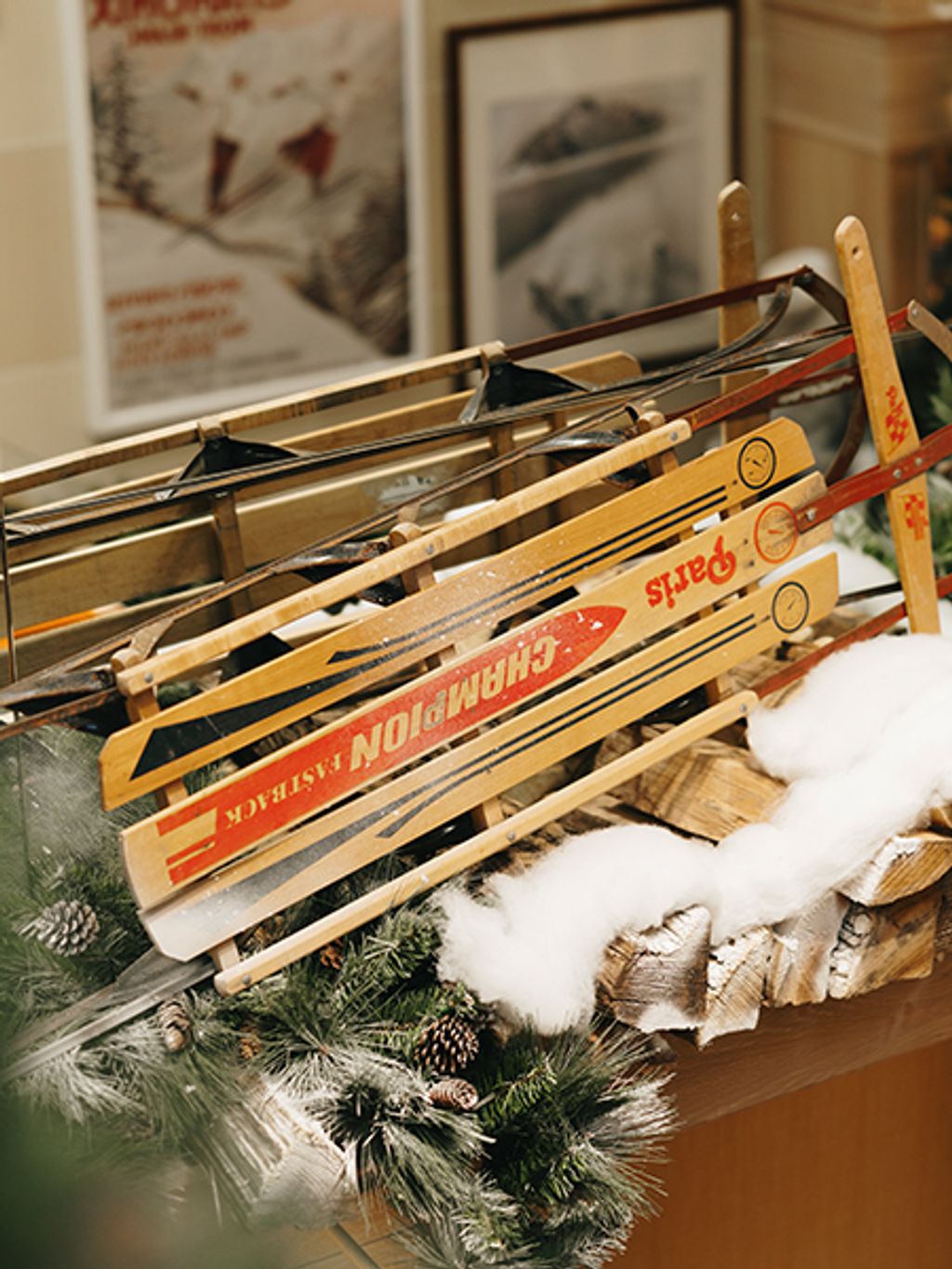 Vintage wooden skis with colorful ‘Champion’ branding are displayed leaning against each other on a snowy, winter-themed setup with faux snow, firewood, and evergreen garland. Framed retro ski posters hang on the wall in the background.