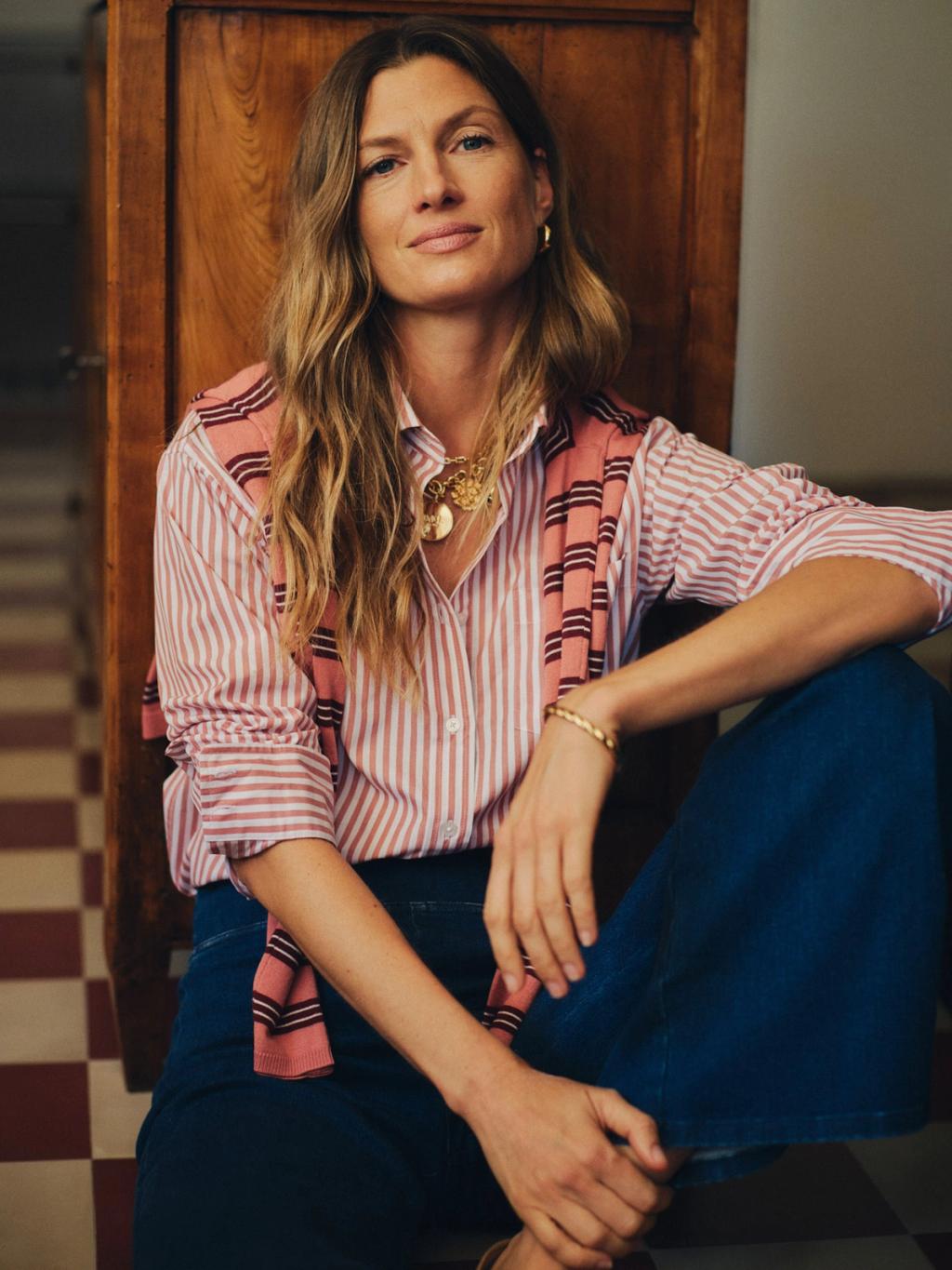 A woman with wavy hair, wearing a pink striped shirt, a sweater over her shoulders, and blue jeans, sits on a tiled floor looking at the camera.