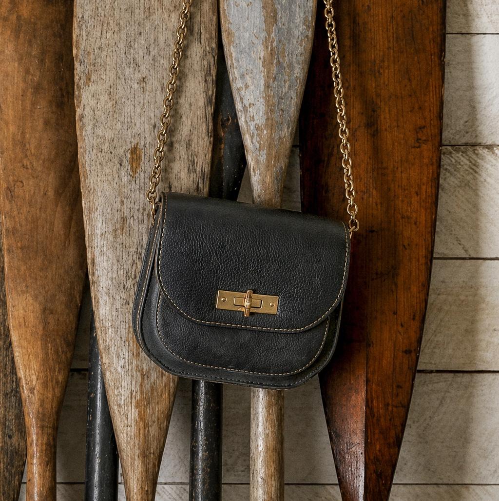 Black leather crossbody bag with a gold chain strap and clasp, hanging against antique wooden paddles and a white plank wall.
