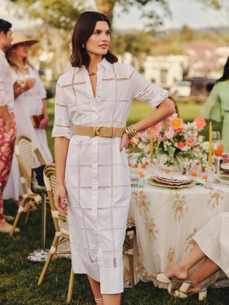 A woman in a white eyelet midi shirt dress and woven belt stands at an outdoor garden party.