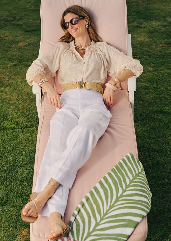 A smiling woman in sunglasses, a patterned blouse, white pants, and a straw belt relaxes on a pink lounge chair on a grassy lawn, next to a palm leaf towel.