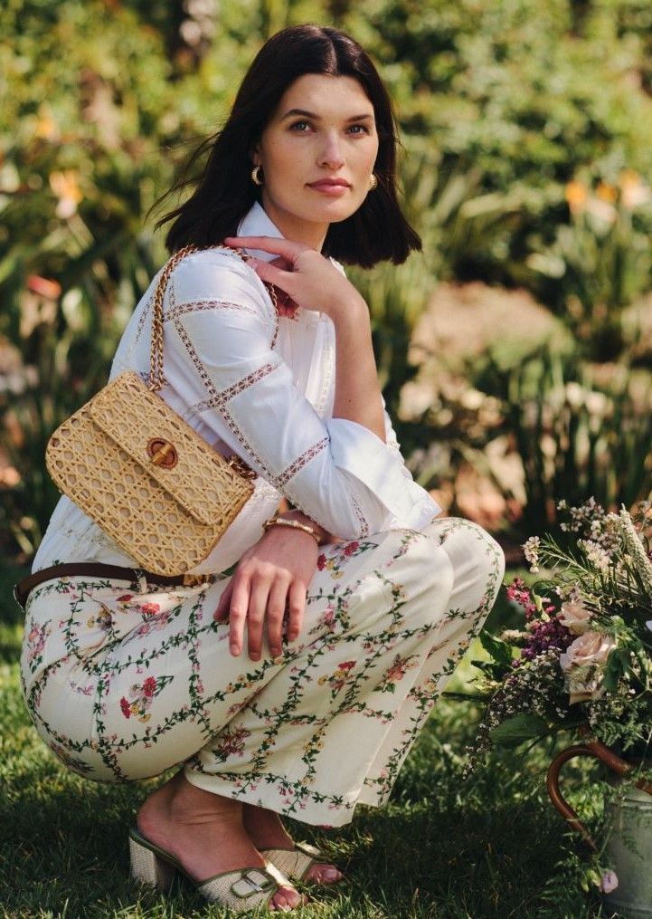 A woman in a white embroidered shirt, floral pants, and straw heels crouches in a garden, holding a rattan bag.