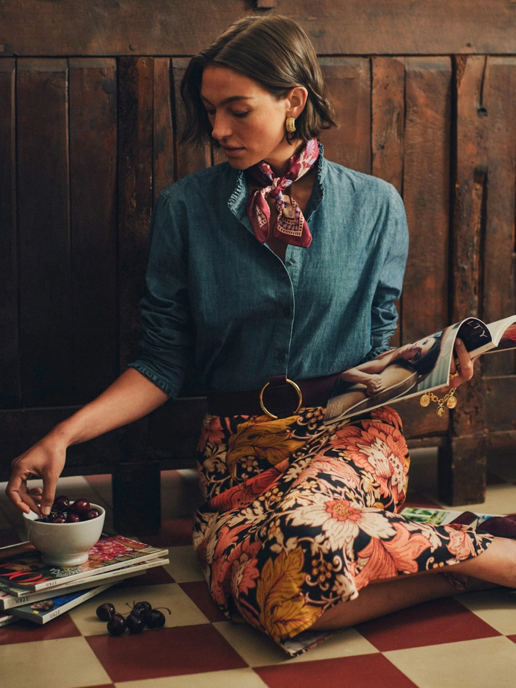A woman in a denim shirt and floral skirt sits on a checkered floor, reaching for cherries from a bowl while holding a magazine.