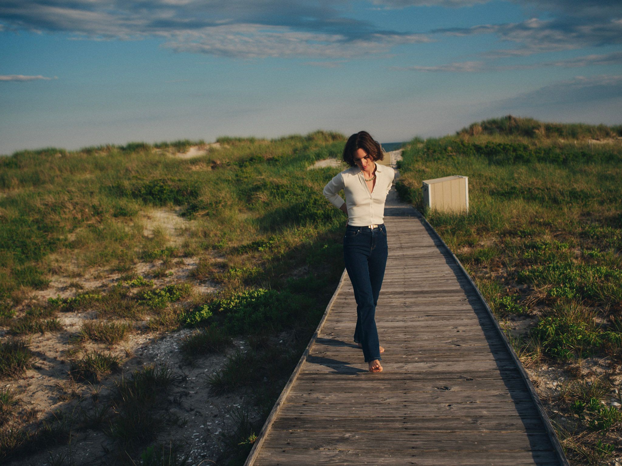 A woman walks on a wooden boardwalk through grassy dunes under a blue sky.