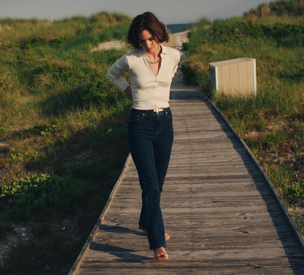 A woman walks on a wooden boardwalk through grassy dunes under a blue sky.