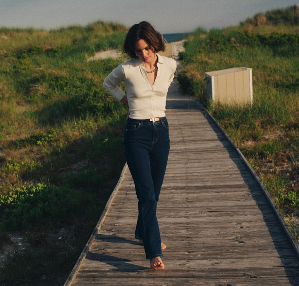 A woman walks along a wooden boardwalk through grassy dunes under a partly cloudy sky.