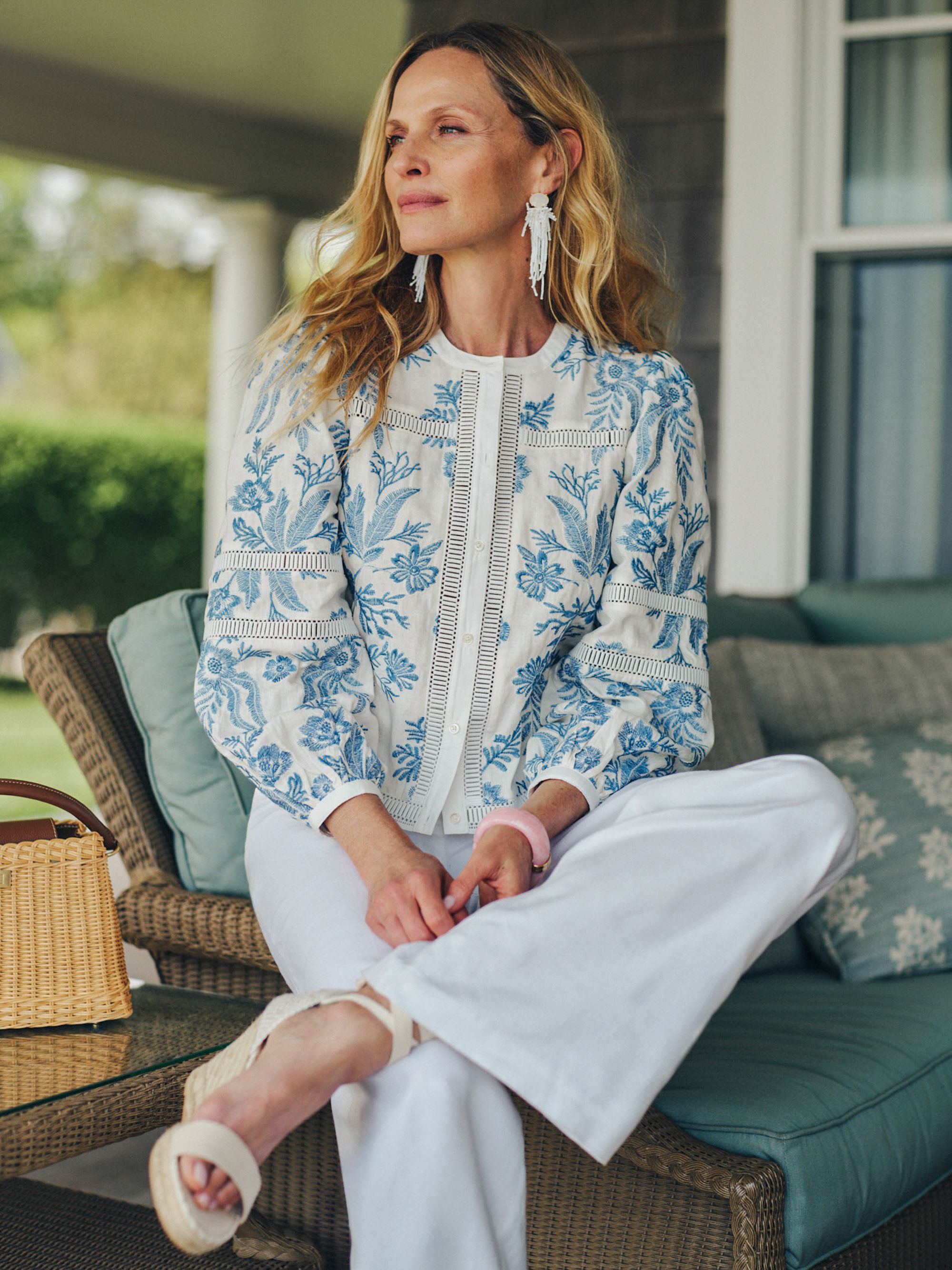 A woman sits on a wicker patio sofa, wearing a white top with blue floral embroidery, white wide-leg pants, white tassel earrings, and a pink bracelet.