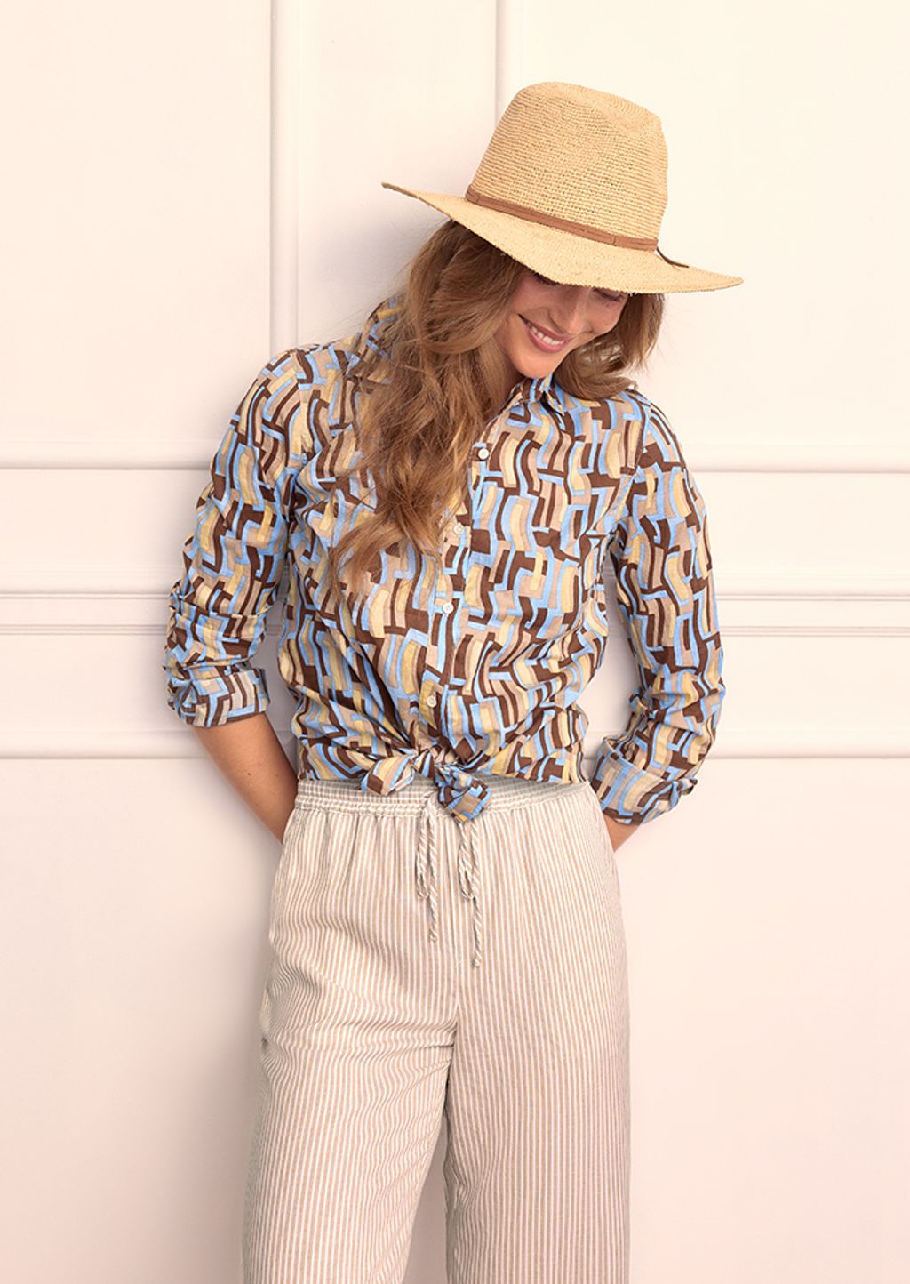 Model wearing a blue and brown geometric shirt with tan and white striped pants and a raffia hat.