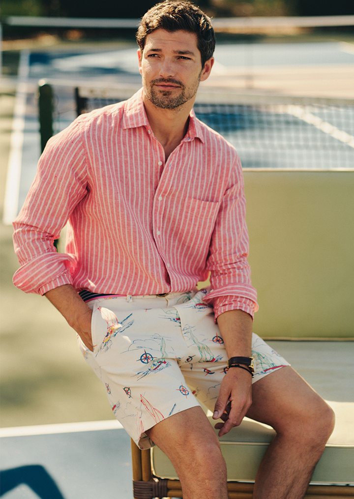 Man walking barefoot on grass in coral-embroidered pink shorts, holding espadrilles.