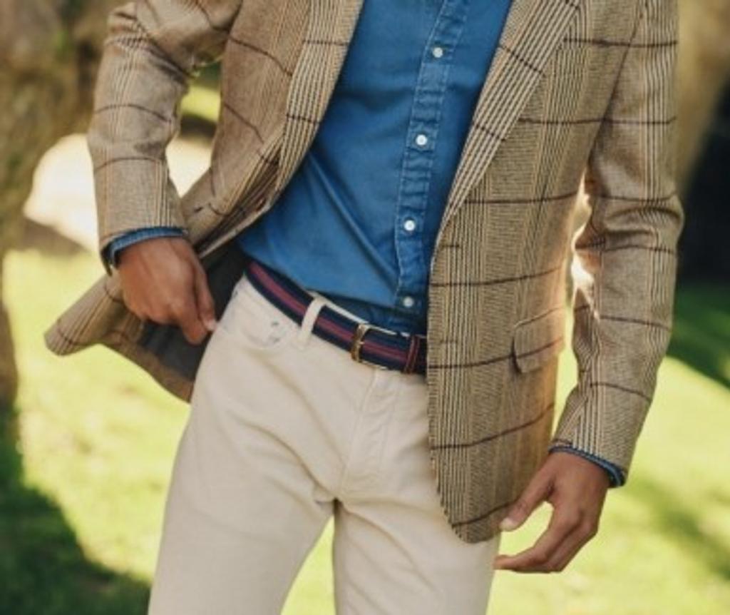 A person wearing a brown plaid blazer, blue shirt, beige pants, and a striped belt.