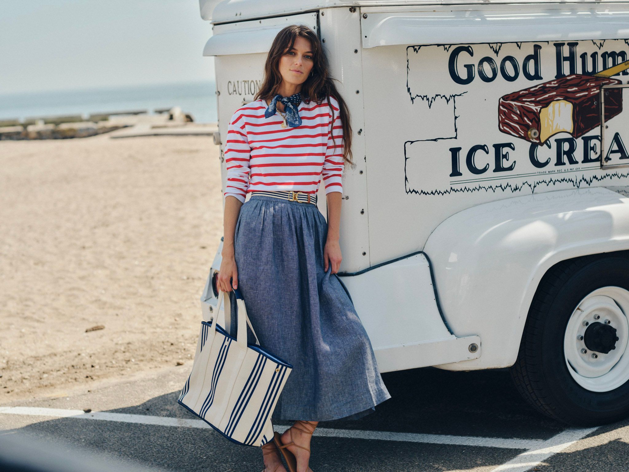 A woman in a red and white striped shirt, blue bandana, and long blue skirt leans against a Good Humor ice cream truck on a sandy beach, holding a striped tote bag.