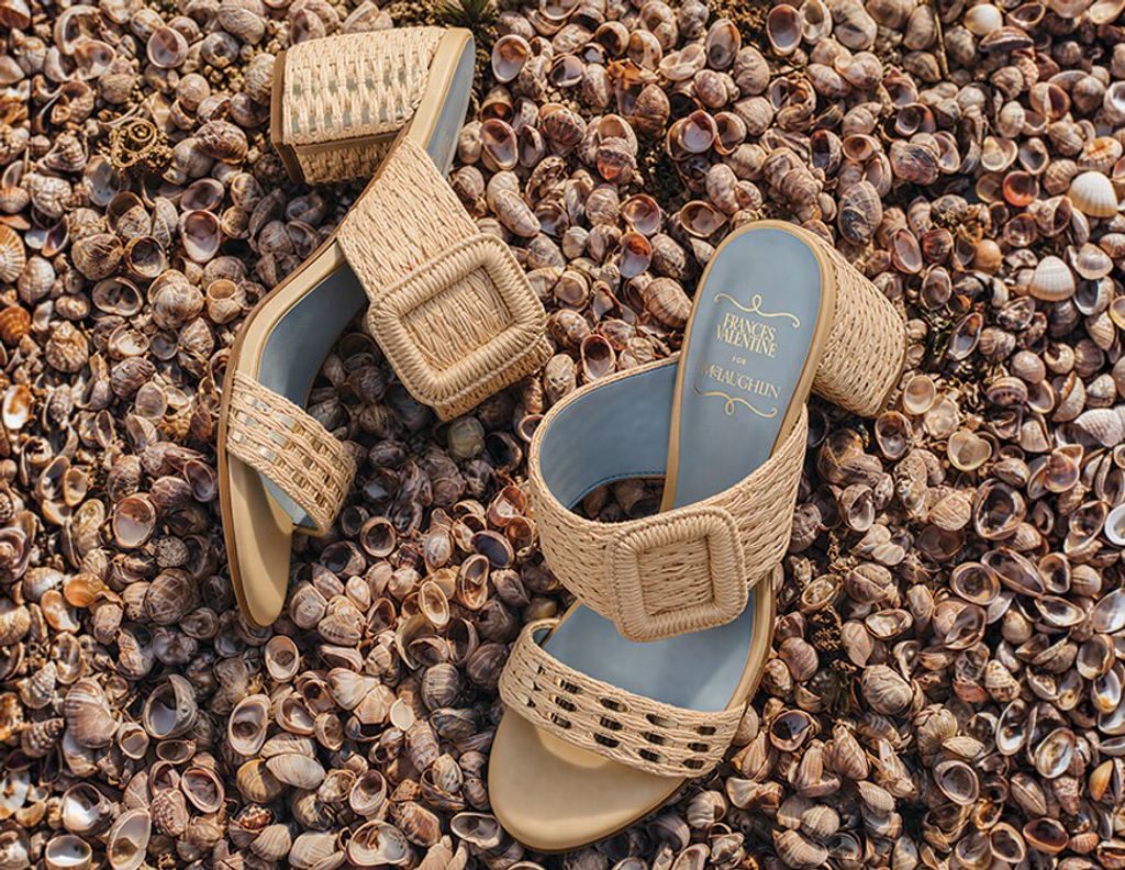 Two woven raffia block-heel sandals with light blue insoles rest on a bed of small seashells.