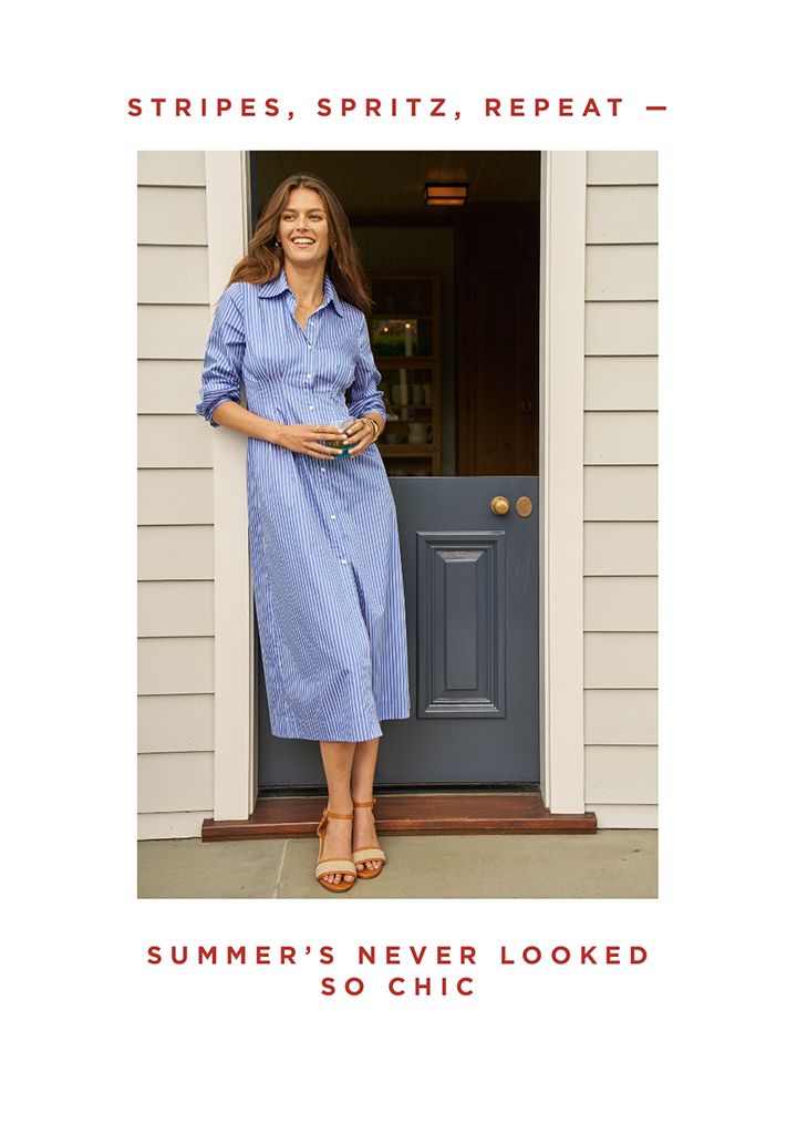 Woman standing outside in a doorway in a blue and white striped dress, with drink in hand.