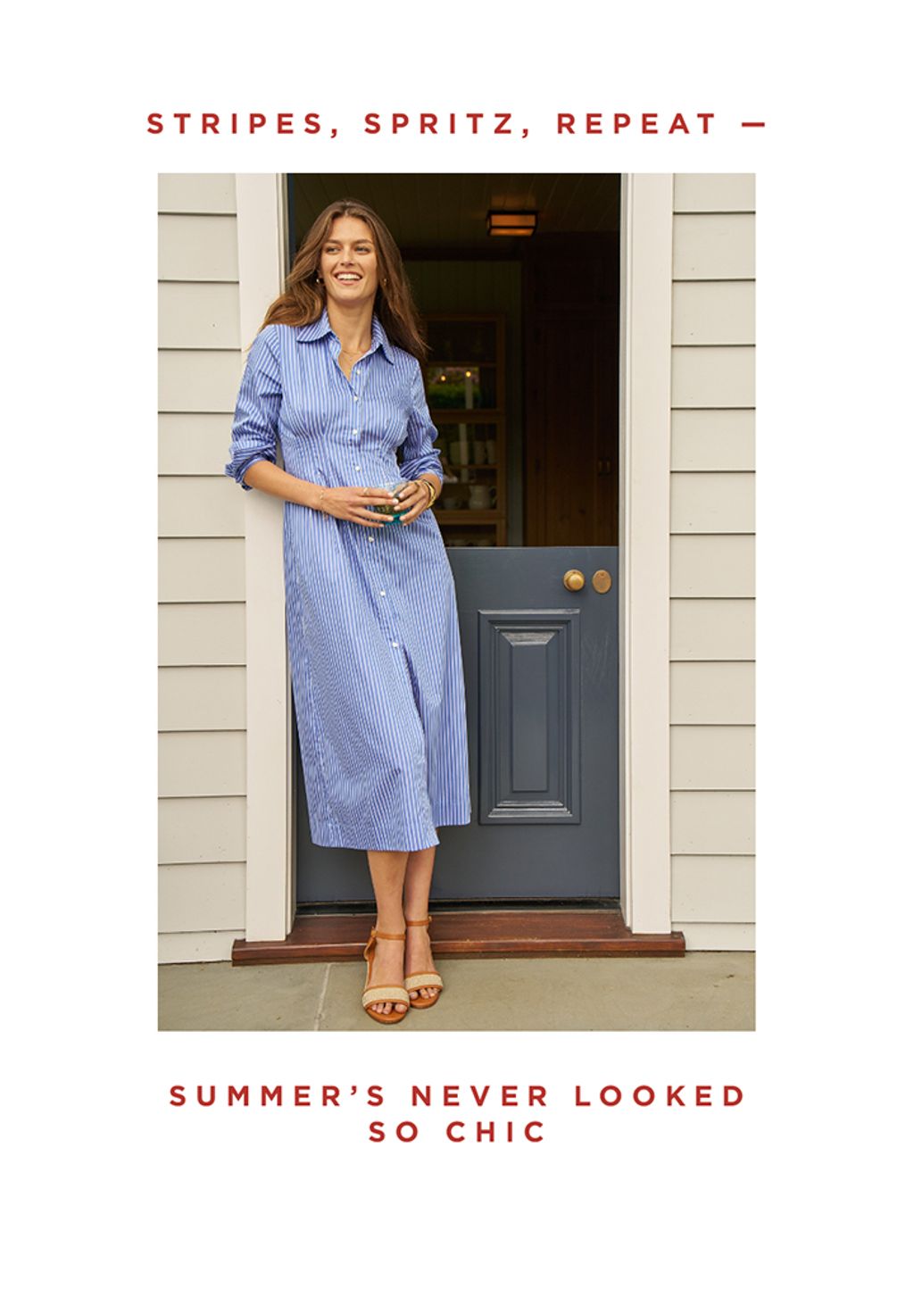 Woman standing outside in a doorway in a blue and white striped dress, with drink in hand.
