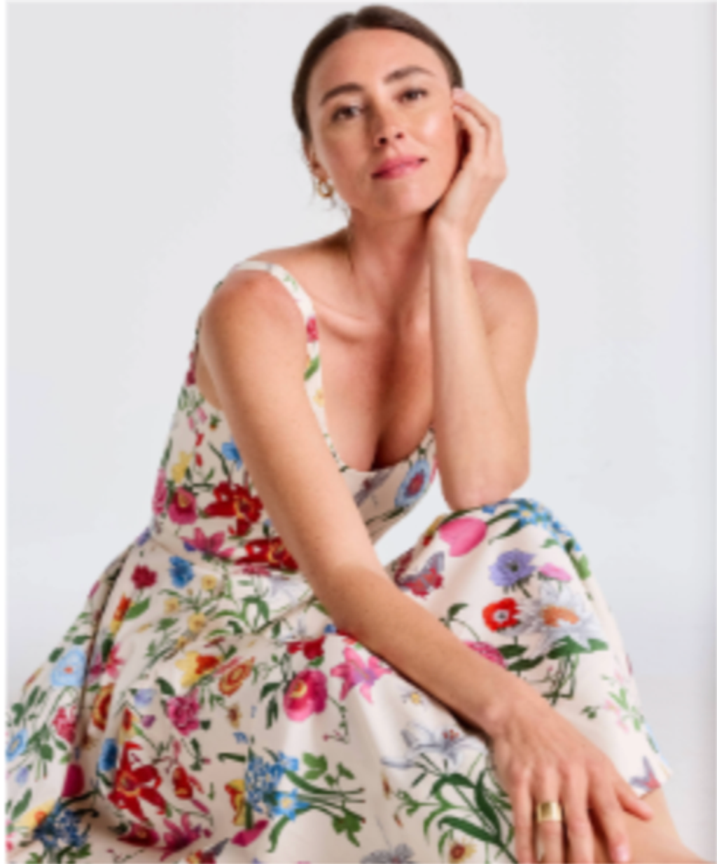 A woman in a colorful floral sundress smiles, resting her chin on her hand.
