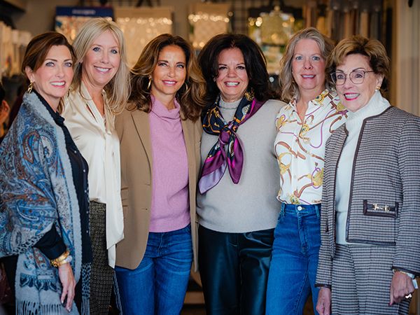 Six women standing together in a line, smiling at the camera in what appears to be a boutique or retail space with soft lighting in the background. From left to right: a woman in a blue patterned cardigan over dark clothing, a woman in a cream blazer, a woman in a tan blazer with a pink turtleneck, a woman in a light gray sweater with a purple and pink patterned scarf and black pants, a woman in a chain-print blouse with blue jeans, and a woman in a gray checkered blazer with glasses.