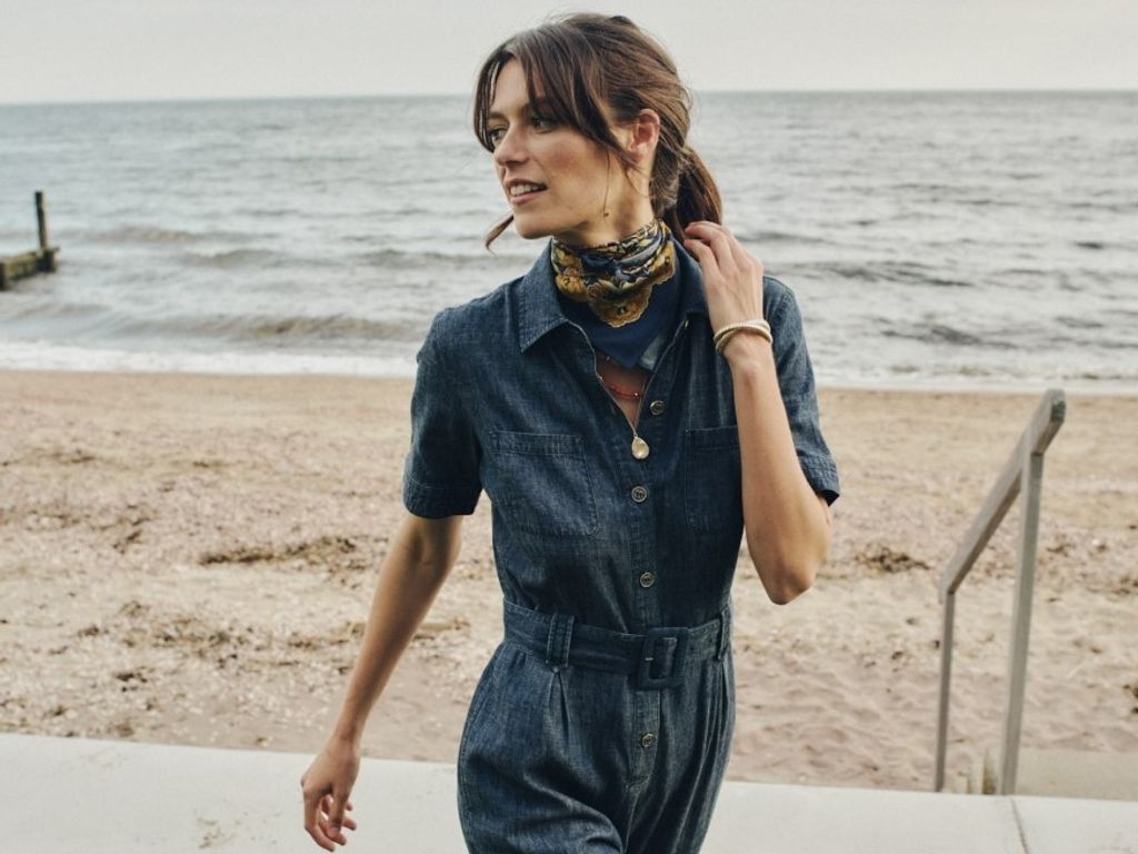 A woman in a denim jumpsuit and patterned scarf stands on a sandy beach by the ocean.