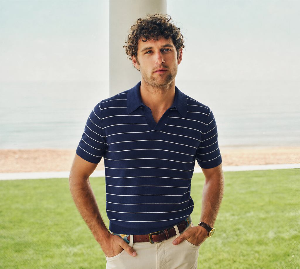 A man in a navy and white striped polo shirt and khaki pants stands on a lawn by a body of water.
