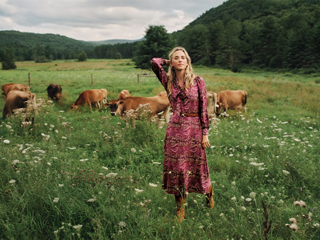 A woman in a pink paisley dress stands in a wildflower field with cows and green hills.