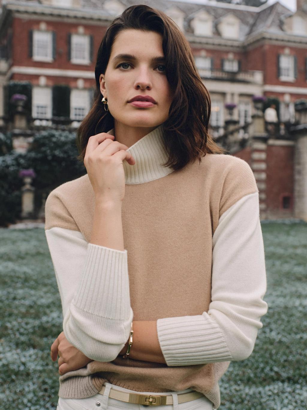 A woman in a camel and cream color-block turtleneck sweater and gold jewelry stands before a large brick building.