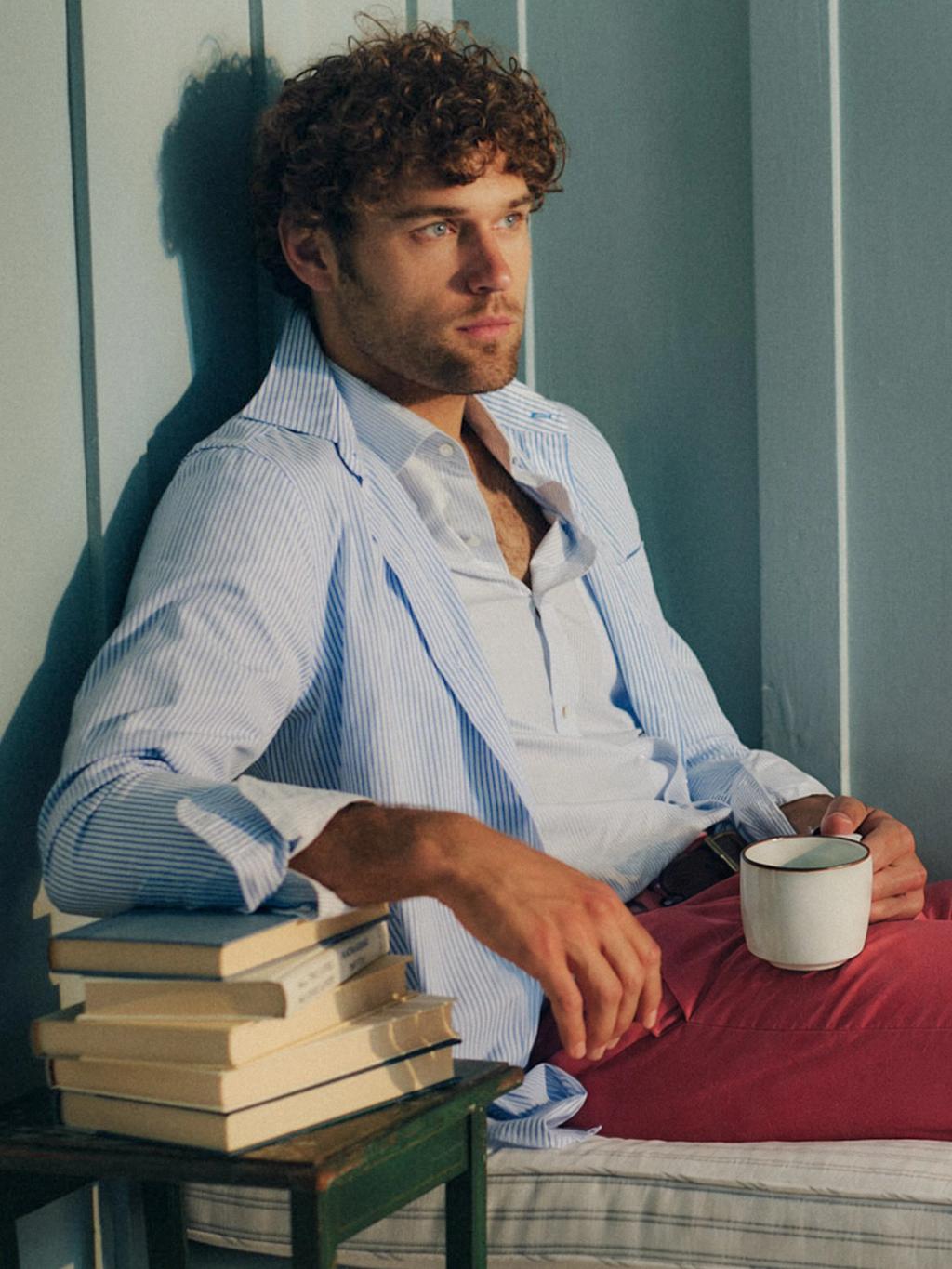A man with curly hair, wearing a striped blue jacket and red pants, sits holding a mug with books stacked next to him.