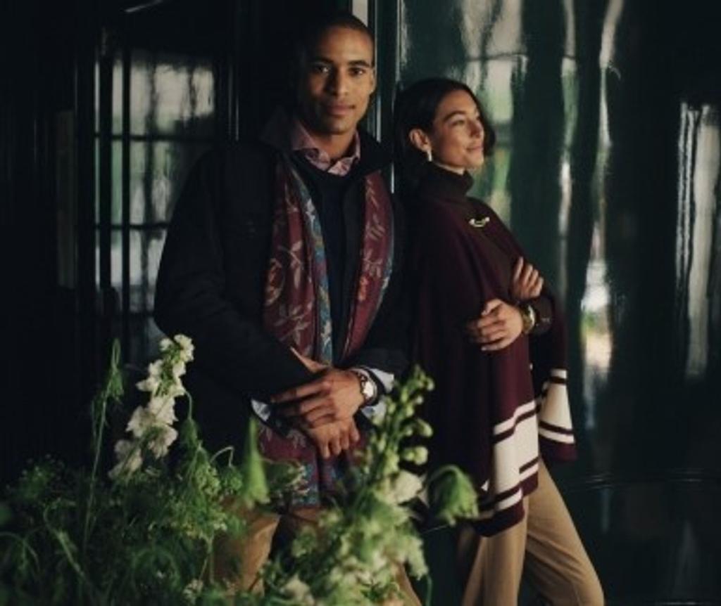 A man in a dark jacket and patterned scarf looks at the camera next to a woman in a burgundy poncho looking right, with flowers in the foreground.