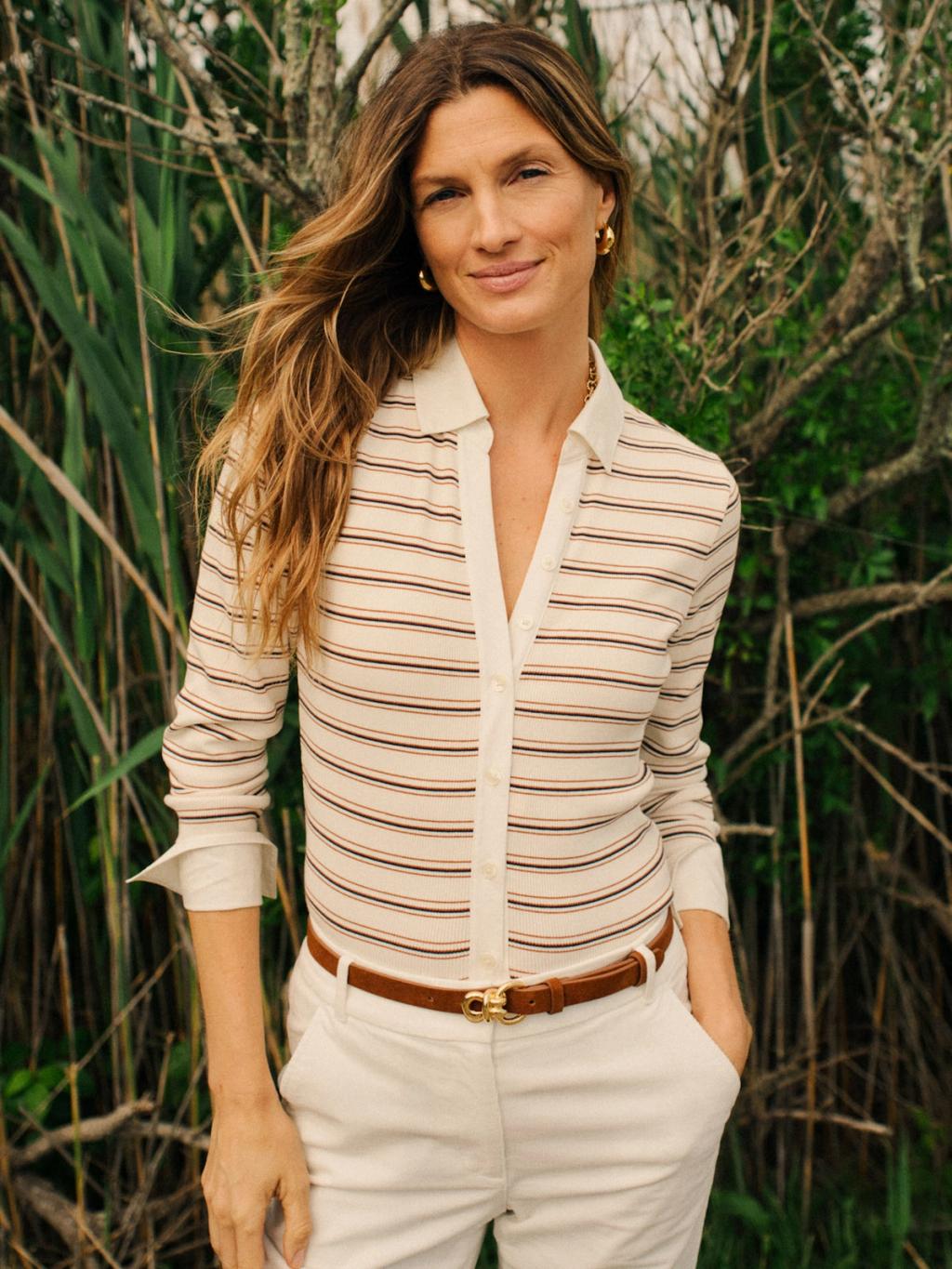 A woman with long wavy brown hair, wearing a cream striped polo shirt, white pants, and a brown belt, smiles slightly outdoors against green foliage.