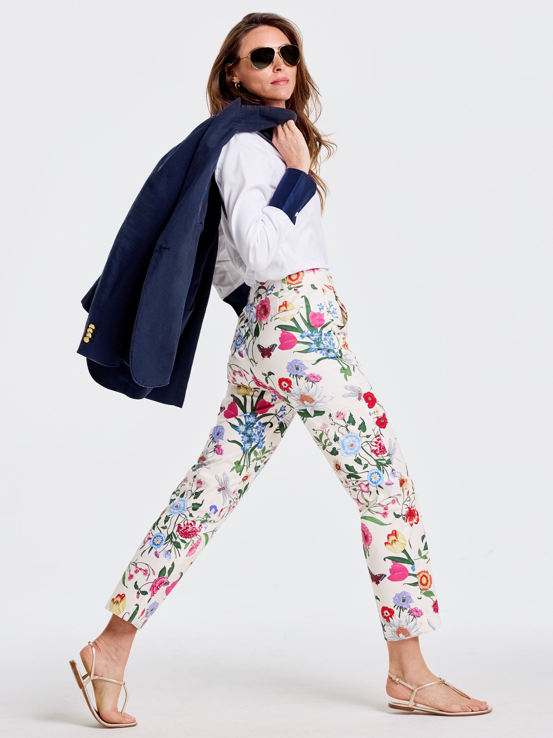 A woman in sunglasses and vibrant floral pants walks, a navy blazer draped over her shoulder.