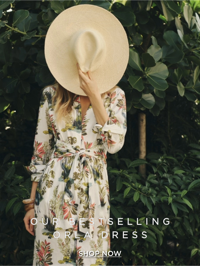 woman standing outside wearing white floral printed shirtdress with sash tie waist and covering her face with straw hat