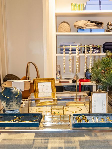 Interior view of a retail clothing store displaying merchandise on racks and shelves, with wrapped gift boxes and various items arranged on a table in the foreground.