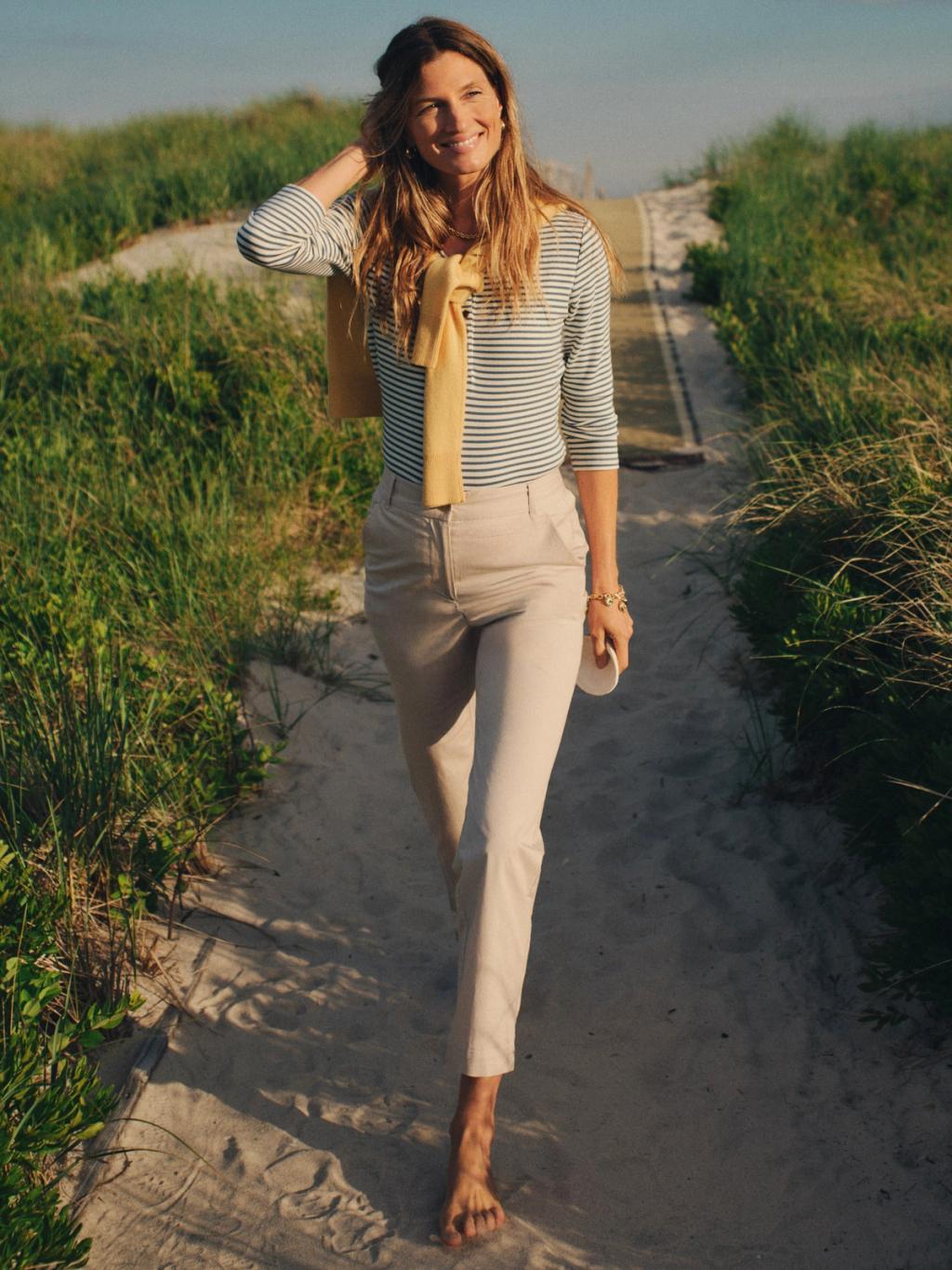 A smiling woman walks barefoot on a sandy path bordered by tall grass, wearing a striped shirt, light pants, and a yellow sweater tied over her shoulders.
