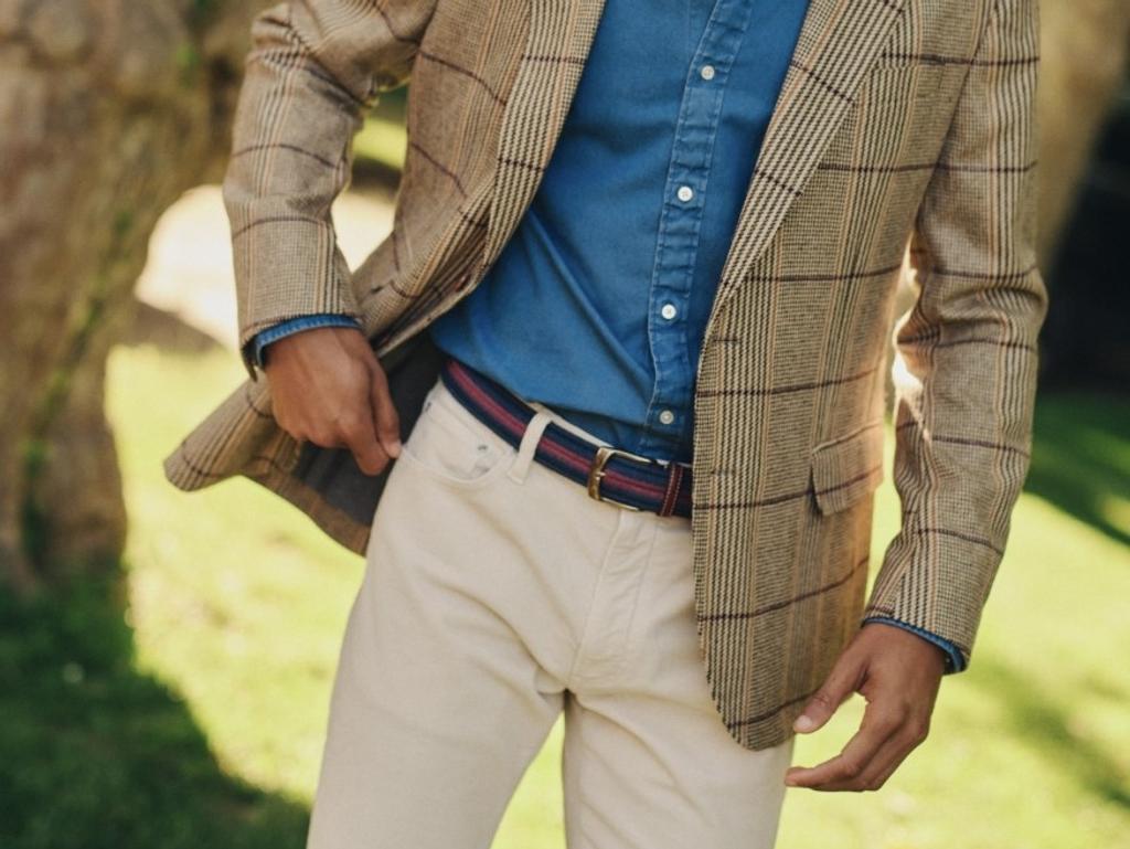 A man wearing a tan plaid blazer, blue shirt, cream pants, and a striped belt, standing outdoors.