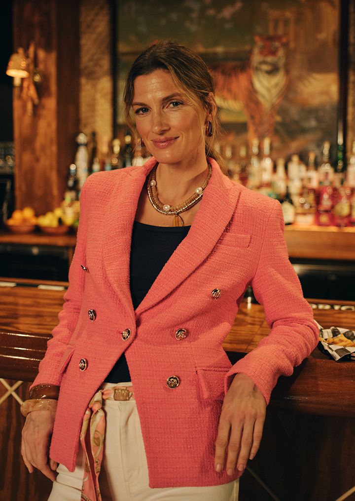 A woman in a coral pink tweed blazer smiles while leaning on a wooden bar.